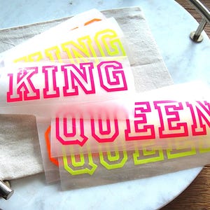 Könnte beinhalten: Eine Sammlung von Aufbügel-Transfers mit den Wörtern KING und QUEEN in einer kräftigen Blockschrift im Varsity-Stil. Die Aufkleber sind in leuchtendem Neon-Pink, hellem Gelb und Orange auf halbtransparentem Trägerpapier dargestellt.