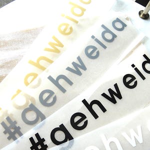 Könnte beinhalten: Vier selbstklebende Vinyl-Aufkleber mit dem Hashtag-Text #gehweida in verschiedenen Farben. Die Aufkleber sind diagonal angeordnet und zeigen goldene, graue, schwarze und weiße Schriftzüge auf einer halbtransparenten Trägerfolie.