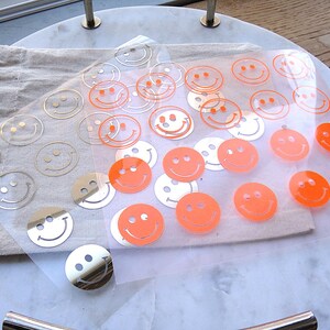 Könnte beinhalten: Zwei transparente Bögen mit mehreren Smiley-Aufklebern. Ein Bogen enthält metallisch silberne Smileys, während der andere Bogen leuchtend neonorangefarbene Smileys zeigt. Die Aufkleber sind in einem Rastermuster auf der Trägerfolie angeordnet.