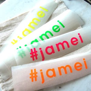 Könnte beinhalten: Vier rechteckige Klebeaufkleber aus Vinyl mit dem Text #jamei in Neongelb, Neongrün, Neonpink und Neonorange. Jeder Aufkleber befindet sich auf einem durchscheinenden Trägerpapier und ist in einer gestapelten Anordnung platziert.
