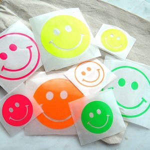 Könnte beinhalten: Eine Sammlung von neonfarbenen Smiley-Aufklebern in verschiedenen Größen. Die Sticker zeigen leuchtend rosa, gelbe, orange und grüne kreisförmige Designs mit zwei Augen und einem geschwungenen Mund auf einzelnen quadratischen Trägerfolien.