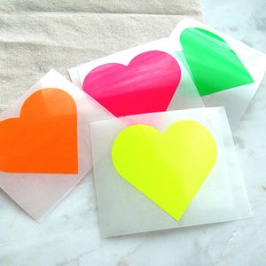Könnte beinhalten: Vier quadratische, durchscheinende Blätter, die jeweils eine einzelne Herzform in leuchtenden Neonfarben wie Orange, Pink, Hellgrün und Gelb zeigen. Die herzförmigen Aufkleber sind in einem überlappenden Muster auf einer Fläche angeordnet.