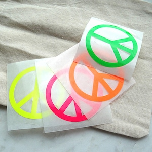 Könnte beinhalten: Ein Set aus vier quadratischen Aufklebern mit einem Friedenszeichen-Symbol in leuchtenden Neonfarben, darunter Grün, Gelb, Pink und Orange. Jeder Aufkleber ist auf einer transparenten Trägerfolie gedruckt und zeigt das kreisförmige Design.