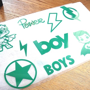 Könnte beinhalten: Ein Bogen mit grünen Vinyl-Aufklebern, die zwei Cartoon-Superhelden, zwei Blitze, einen Stern in einem Kreis sowie den Text "Power", "boy" und "BOYS" zeigen. Die Motive sind aus einem Stück grünem Klebematerial auf einer weißen Trägerfolie geschnitten.