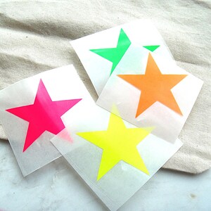 Könnte beinhalten: Vier einzelne quadratische Bögen aus durchscheinendem Transferpapier, die jeweils einen fünfzackigen Stern in einer leuchtenden Neonfarbe zeigen. Die Sterne sind in leuchtendem Pink, Limettengrün, Orange und Gelb auf einer flachen Oberfläche angeordnet.