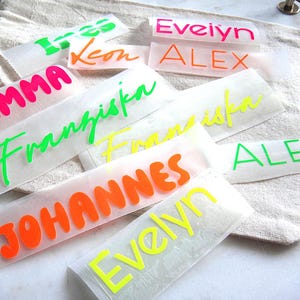 Op de afbeelding: Een verzameling rechthoekige zelfklevende vinylstickers met diverse namen in neonkleuren, waaronder roze, oranje, groen en geel. De namen die op het transparante transferpapier zijn gedrukt, zijn onder andere Ines, Leon, Evelyn, Alex, Emma, Franziska en Johannes.