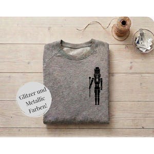 Könnte beinhalten: Ein graumeliertes Sweatshirt mit Rundhalsausschnitt und einem schwarzen Nussknacker-Motiv auf der Brust. Ein weißes, kreisförmiges Etikett auf der linken Seite zeigt den schwarzen Text Glitzer und Metallic Farben, der auf Glitzer- und Metallic-Farben hinweist.