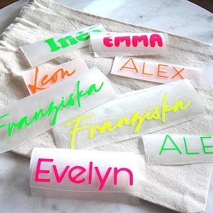 Könnte beinhalten: Eine Sammlung personalisierter Vinyl-Namensaufkleber in verschiedenen Neonfarben wie Pink, Grün, Orange und Gelb. Die Namen Ines, Emma, Leon, Alex, Franziska und Evelyn sind in verschiedenen Schriftarten auf einzelnen Transferfolien gedruckt.