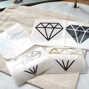 Könnte beinhalten: Fünf quadratische Aufkleber mit einem Diamant-Umriss-Design. Die Sammlung umfasst eine weiße, eine schwarze, eine vollflächig schwarze, eine silberne und eine goldene Diamantgrafik, die jeweils auf einer transparenten Trägerfolie präsentiert werden.