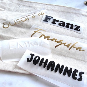 Op de afbeelding: Een verzameling opstrijkbare vinylstickers met de namen Susanne, Franz, Franziska, EMMA en JOHANNES in verschillende lettertypes en kleuren, waaronder goud, zwart en wit, gerangschikt op een beige stoffen tasje voor toepassing.