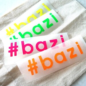 Könnte beinhalten: Vier rechteckige Klebeaufkleber mit dem Hashtag-Text #bazi in einer serifenlosen Kleinbuchstabenschrift. Die Aufkleber sind in den Farben Neongelb, Neongrün, Neonpink und Neonorange gehalten und auf einer durchscheinenden Trägerfolie.
