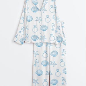 Op de afbeelding: Witte pyjamaset met een blauw patroon van zeeschelpen, zeesterren, champagneglazen en diamanten ringen. De top met lange mouwen heeft een knoopsluiting en een reverskraag. De bijpassende broek heeft een elastische tailleband.