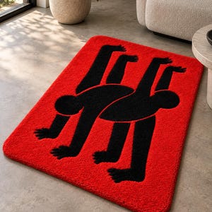 May include: A rectangular area rug featuring a vibrant red background with a large, black, stylized graphic of two interconnected figures. The figures are depicted in a continuous, abstract form with multiple limbs, creating a bold contrast against the red.