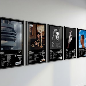 May include: A series of framed album art prints featuring Drake. Each black-framed print displays album art, tracklists, and artist information. The images include portraits and album covers, creating a cohesive music-themed wall display.
