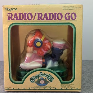 Peut inclure: Un jouet radio AM Cabbage Patch Kids dans son emballage en carton jaune d'origine. Le jouet représente un personnage aux cheveux roux portant un casque bleu, un haut rose et un pantalon bleu, posé sur une base vert foncé. Texte sur la boîte.