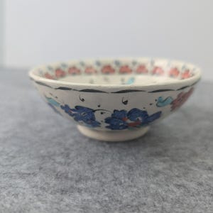 May include: A small, shallow ceramic bowl with a white base, featuring hand-painted floral motifs in shades of blue, red, and black. The exterior rim is decorated with a repeating pattern of small red petals, while the body displays larger blue flowers.