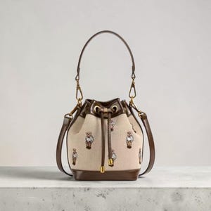 Può includere: Una borsa a secchiello in tela beige con un motivo ricamato di piccoli orsetti che indossano maglioni. La borsa presenta finiture in pelle marrone scuro, chiusura a coulisse con dettagli in metallo dorato, un manico e una tracolla rimovibile.
