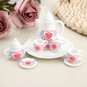 Puede incluir: Un juego de té en miniatura de cerámica blanca con un patrón repetido de corazones rosas con detalles en azul y naranja. El conjunto incluye una tetera con tapa, un azucarero con tapa, una jarra de leche, dos tazas, dos platillos y un pequeño plato decorativo.