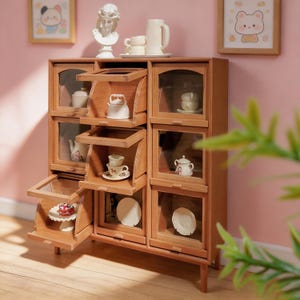 Puede incluir: Un armario de madera en miniatura con nueve compartimentos y puertas de cristal abatibles. El armario exhibe pequeños juegos de té de cerámica, platos y un pastel en un soporte. Un busto blanco y una jarra descansan sobre la parte superior.