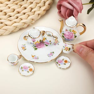 Może przedstawiać: Miniaturowy zestaw do herbaty z białej porcelany z delikatnymi wzorami różowych róż i żółtych kwiatów, wykończony złotymi brzegami. Zestaw zawiera imbryk, dzbanek na mleko, cukiernicę, małe filiżanki oraz spodki z motywem kwiatowym.