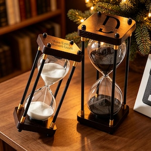 May include: Two hourglasses with dark wood frames, gold accents, and black support rods. One contains white sand and is engraved with "Make Every Moment Count," while the other contains black sand and is engraved with the name "DANGELMAYR" on the top.