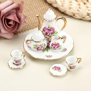 Peut inclure: Un service à thé miniature en porcelaine comprenant une théière, un sucrier, un pot à lait et deux tasses avec soucoupes assorties. Chaque pièce blanche est ornée d'un motif floral rose, de feuilles vertes et d'un liseré doré sur les bords.