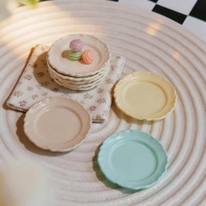 以下が含まれることがあります： クリーム色、黄色、水色のパステルカラーの縁が波打ったミニチュア陶器皿のセット。クリーム色の皿の山の上には、ピンク、緑、オレンジの小さなカラフルなマカロンが乗っており、花柄のナプキンの上に置かれています。