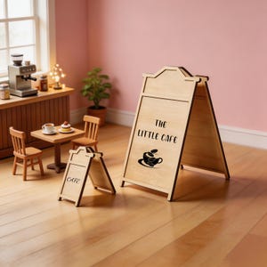 May include: Two light-colored wooden A-frame signs for a miniature cafe. The larger sign features the text THE LITTLE CAFE and a graphic of a coffee cup with beans. The smaller sign displays the word CAFE. Both are made of smooth, light wood.