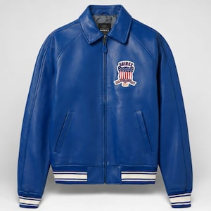 Könnte beinhalten: Eine leuchtend blaue Lederjacke mit durchgehendem Frontreißverschluss, klassischem Kragen sowie gerippten Bündchen und Saum mit weißen Streifen. Auf der linken Brust befindet sich ein gestickter Aufnäher mit dem Text AVIREX über einem US-Flaggenschild.