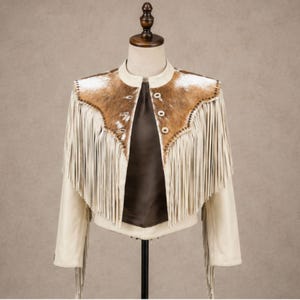 Puede incluir: Una chaqueta de color crema con un canesú de piel de vaca marrón y blanca en el pecho y los hombros. La prenda incluye flecos largos y finos a lo largo del canesú delantero y las mangas, un cuello mao y tres botones circulares de color claro.