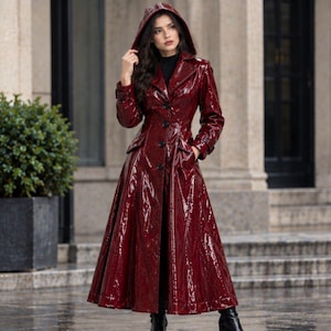 May include: A long, glossy, dark red trench coat made of a patent leather-like material. The coat features a hood, a wide collar, long sleeves with buckled cuffs, and a row of black buttons down the front. It has a fitted waist and a flared skirt.