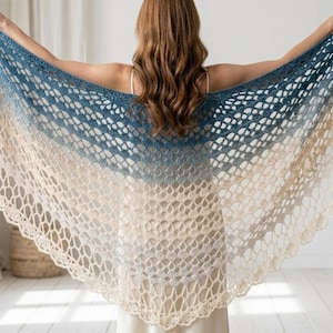 Puede incluir: Un gran chal triangular de ganchillo con un intrincado patrón calado. El diseño muestra un degradado de color que transita desde un azul cerceta profundo en el borde superior hasta un blanco crema suave en el borde inferior festoneado.