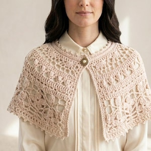 Könnte beinhalten: Ein hellbeiges Häkel-Capelet mit einem komplizierten Blumenmuster und durchbrochenen Details. Das Kleidungsstück wird am Halsausschnitt mit einem einzelnen runden Knopf befestigt, der einen perlmuttartigen Akzent und einen strukturierten Metallrand hat.