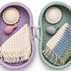 May include: Two oval crochet baskets, one lavender and one sage green, each featuring side handles. Inside the lavender basket is a cream circular mat, a pink rattle, and a cream fringed blanket. The green basket contains a cream mat, a gray fish-shaped rattle, and a blue, green, and cream striped fringed blanket.