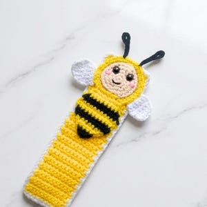 May include: A handmade crochet bookmark in the shape of a bee. The bookmark features a yellow and black striped body, a yellow head with a smiling face, white wings, and black antennae. The bookmark is a cute and whimsical accessory.