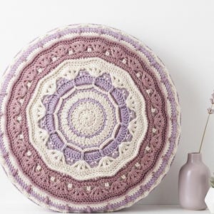 May include: A circular, crocheted cushion in cream, lavender, and mauve. The cushion has a detailed, mandala-style design with intricate stitch patterns. A small vase with a sprig of lavender and a succulent in a white pot are in the background.