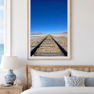 May include: A framed photograph of a railway track receding into the distance under a clear, bright blue sky. The track is flanked by a barren landscape with distant mountains. The photograph is displayed above a bed with blue and white bedding.