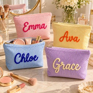 Puede incluir: Cuatro neceseres con cremallera hechos de tela seersucker en rosa, amarillo, azul y morado. Cada neceser presenta un nombre bordado en un color contrastante: Emma en rosa, Ava en naranja, Chloe en azul y Grace en crema.