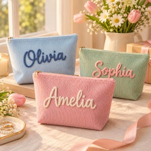 Könnte beinhalten: Drei Reißverschlusstaschen aus Kord in Hellblau, Salbeigrün und zartem Rosa. Jede Tasche ist mit einem in Schreibschrift gestickten Namen versehen: Olivia in Dunkelblau, Sophia in Rosa und Amelia in Weiß. Die Taschen haben goldfarbene Reißverschlüsse.