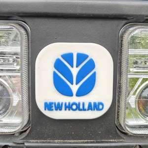 Op de afbeelding: Een wit en blauw New Holland logo tussen twee koplampen van een voertuig. Het logo toont een blauw bladontwerp boven de woorden "NEW HOLLAND" in blauw, tegen een witte achtergrond.