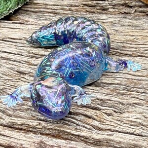May include: A translucent resin lizard figurine featuring a deep blue and purple colour palette with embedded iridescent glitter and small metallic shards. The creature has a segmented tail, four splayed feet, and a smooth, glossy finish.