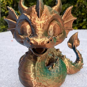 Puede incluir: Una figura de dragón impresa en 3D con un acabado metálico que cambia de color entre cobre y verde. La criatura tiene ojos grandes, cuernos pequeños, orejas extendidas y una cola enroscada. La superficie muestra capas horizontales visibles.