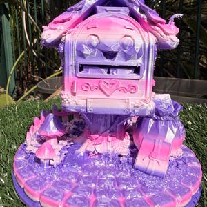 May include: A 3D printed post box structure featuring a gradient of purple and pink colours. The post box has a heart-shaped emblem, a roof with textured details, and sits on a base with small mushroom figures, a gift box, and a sack.