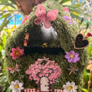May include: A whimsical fairy house with a moss-covered roof, adorned with pink and white flowers, butterflies, and a miniature tree with a door. A heart-shaped sign reads "Fairy Mail".