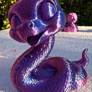 May include: A 3D printed snake figurine with a glossy, iridescent purple and blue finish. The snake has a large head, prominent eyes, and an open mouth, with a coiled body featuring visible layer lines from the printing process.