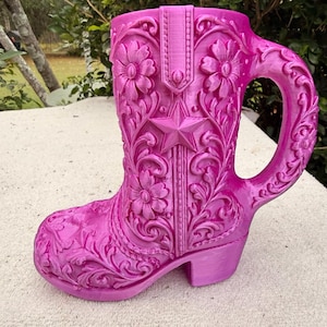 May include: A bright pink mug shaped like a cowboy boot, featuring intricate embossed floral patterns, scrolling vine details, and a prominent five-pointed star on the front shaft. The handle is integrated into the back of the boot.
