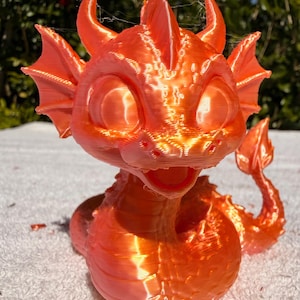 Puede incluir: Una figura de dragón impresa en 3D de color naranja brillante y lustroso. La criatura presenta ojos grandes y redondeados, orejas puntiagudas, una cresta central en la cabeza y alas desplegadas. Su cuerpo está enroscado con una textura visible.