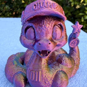 May include: A 3D printed iridescent purple and gold snake figurine wearing a baseball cap with the word CHAMP embossed on the front and the number 1011 on its chest. The snake has large eyes, an open mouth, and a star-shaped rattle on its tail.