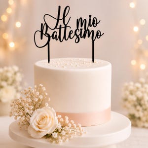 Può includere: Una torta bianca con un nastro rosa, sormontata da un cake topper nero con la scritta "Il mio Battesimo". La torta è decorata con una rosa color crema e piccoli fiori bianchi. La torta è su un vassoio bianco.