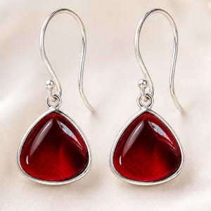 May include: A pair of silver drop earrings featuring teardrop-shaped, deep red stones. Each earring has a smooth, polished silver hook and a bezel setting that secures the vibrant, translucent red gemstone, creating a balanced and symmetrical design.